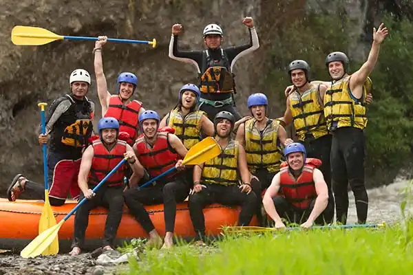 A group of nine happy rafters in yellow and red life vests posing with their paddles on the riverbank