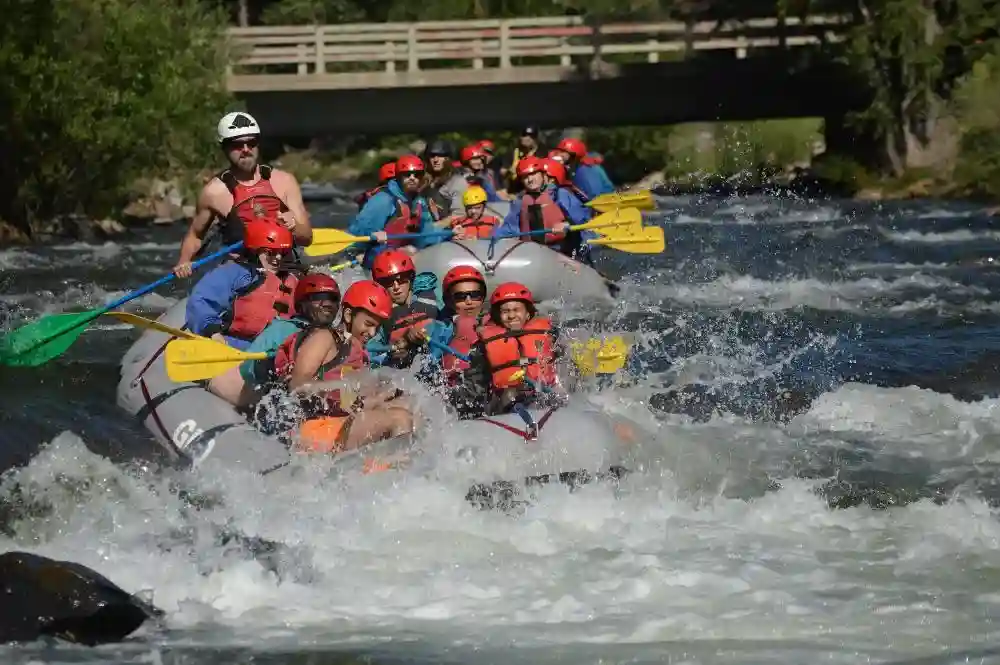 A large grey raft filled with people in red helmets paddling through splashing white water under a wooden bridge